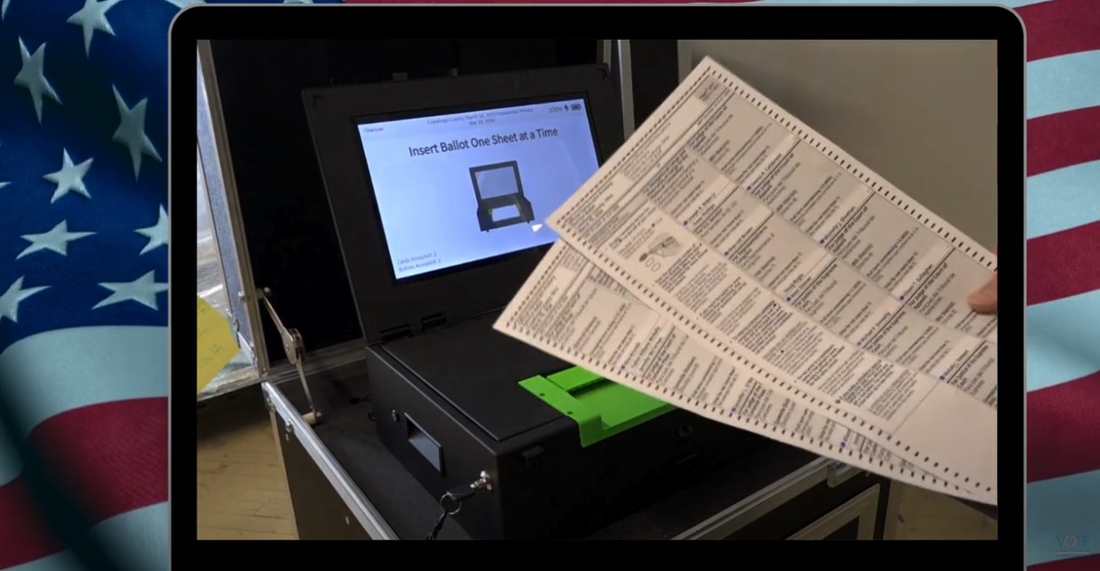 An image of the ClearCast Go ballot scanner and a voter holding their two-page ballot in front of the machine, preparing to insert their ballot into the scanner